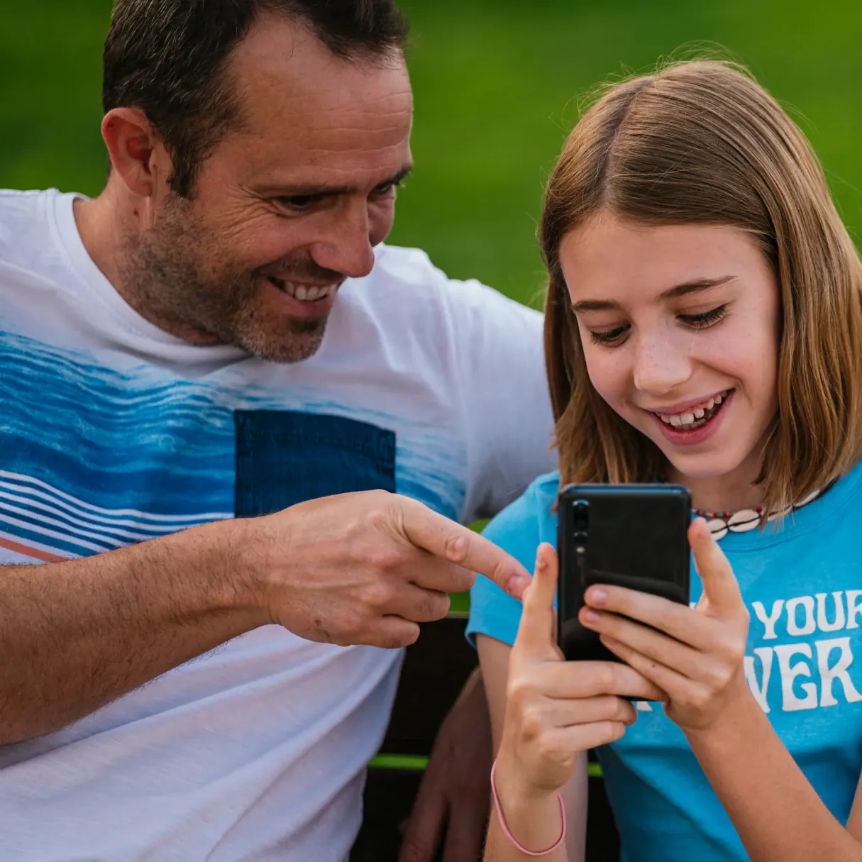 Dad pointing at his daughter's phone screen as they look at it together outside
